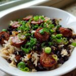 A white bowl filled with One-Pot Black Beans and Rice with Sausage, topped with sliced smoked sausage and green onions.