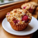 A close-up of a freshly baked Raspberry Streusel Muffins topped with golden brown streusel and bright red raspberries.