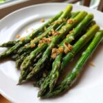 Bright green Roasted Asparagus with Garlic and Lemon piled on a white plate, glistening with oil.