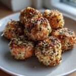A close-up of several golden-brown Salmon Rice Bites coated in mixed sesame seeds on a light gray plate.