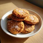 A stack of golden brown Salted Honey Cookies topped with visible flakes of sea salt, served on a light plate.
