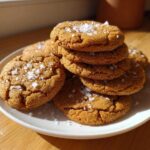 A stack of golden brown Salted Honey Cookies topped generously with flaky sea salt, resting on a white plate.