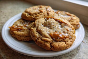 Four freshly baked Sea Salt Caramel Cookies topped with gooey caramel and flaky sea salt on a white plate.