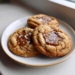 Three freshly baked Sea Salt Caramel Cookies topped with gooey caramel centers and flaky sea salt.