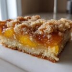 A close-up, side-view of a thick Peach Crumb Bars square with a buttery crust, bright orange peach filling, and crumbly topping.