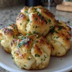 A close-up photo of several soft Garlic Knots piled on a white plate, glistening with butter and topped with fresh green parsley.