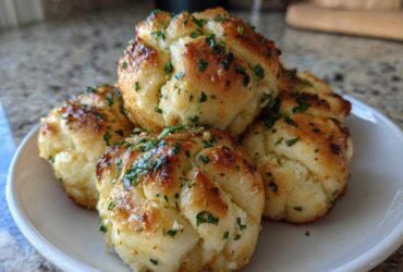 A close-up photo of several soft Garlic Knots piled on a white plate, glistening with butter and topped with fresh green parsley.