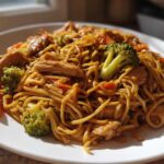 A close-up, appetizing serving of Stir-Fried Noodles mixed with sliced chicken, bright green broccoli florets, and carrots on a white plate.