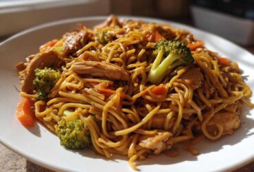 A close-up, appetizing serving of Stir-Fried Noodles mixed with sliced chicken, bright green broccoli florets, and carrots on a white plate.
