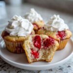 A close-up of several Strawberry Shortcake Muffins topped with whipped cream, one muffin is bitten into showing the strawberry filling.