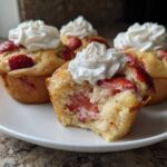 A close-up of several Strawberry Shortcake Muffins topped with whipped cream, one muffin is broken open showing the strawberry filling.
