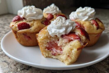 A close-up of several Strawberry Shortcake Muffins topped with whipped cream, one muffin is broken open showing the strawberry filling.