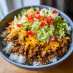 Close-up of a bowl of Taco Rice topped with seasoned ground beef, melted cheese, shredded lettuce, and diced tomatoes.