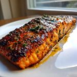 A perfectly cooked, glazed Texas Roadhouse Salmon Copycat fillet topped with herbs, resting in its juices on a white plate.