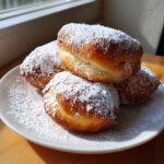 A stack of four golden-brown Vanilla French Beignets generously dusted with powdered sugar on a white plate.